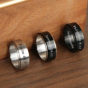 May include: Three rings with engraved text. One ring is silver with "Psalm 46:5", another is black with "Jeremiah 29:11", and the third is black with "Philippians 4:13 Just Breathe". The rings are on a wooden surface.