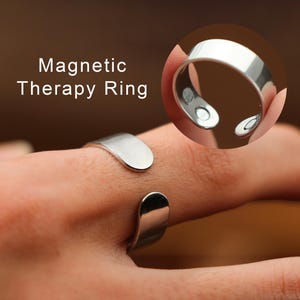 May include: A silver magnetic therapy ring. The ring is open-ended with a smooth, polished finish. The text "Magnetic Therapy Ring" is displayed in white. The ring has two small magnets visible on the inside.
