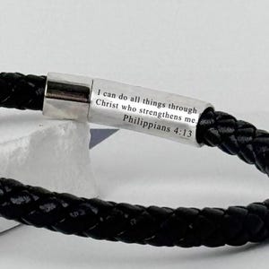 May include: Black braided leather bracelet with a silver clasp. The clasp is engraved with the text "I can do all things through Christ who strengthens me. Philippians 4:13". The bracelet is a religious accessory.