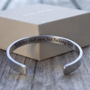 May include: A silver-toned metal cuff bracelet with an open design. The interior surface of the bracelet is engraved with the black text, "This too shall pass, but fucking hell." The bracelet has a smooth, polished finish and a rounded band shape.