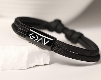 Op de afbeelding: Zwart geweven armband met een rechthoekige zwarte sluiting. De sluiting heeft de witte letters G>AV. De armband ligt op een wit, getextureerd oppervlak. De armband is gemaakt van een dik, duurzaam materiaal.