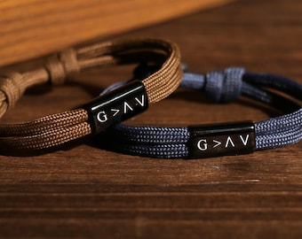 Op de afbeelding: Twee verstelbare armbanden met zwarte sluitingen. Eén is bruin, de andere blauw. Elke sluiting heeft de tekst "G > V" in het wit. De armbanden zijn gemaakt van gevlochten koord en liggen op een houten oppervlak.