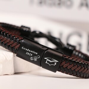 May include: A brown and black braided leather bracelet with a black clasp. The clasp is engraved with "Graduation 2025" and a graduation cap and scroll icon. A fashionable accessory.