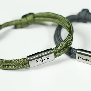 May include: Two braided rope bracelets with personalized metal tags. The green bracelet has an anchor and the letters "A" and "K" engraved on the tag. The gray bracelet has the name "Thomas" engraved on the tag.