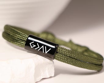 Op de afbeelding: Een groene paracord armband met een zwarte metalen schuifregelaar met het christelijke symbool "G.O.A.T." in wit.