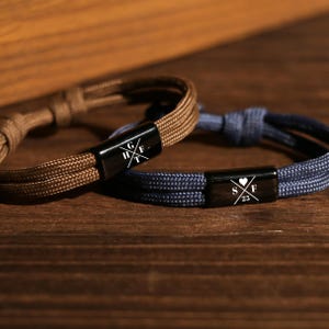 May include: Two cord bracelets, one brown and one navy blue, each featuring a black rectangular metal centrepiece. The brown bracelet is engraved with the letters G, H, F, and T in a cross pattern. The navy bracelet features S, F, 25, and a heart.