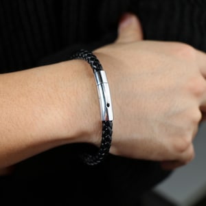 Puede incluir: Un brazalete de cuero negro con un cierre de plata. El brazalete est&aacute; trenzado y tiene un dise&ntilde;o sencillo.