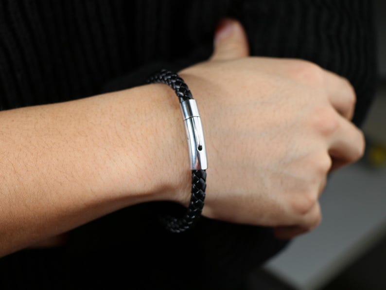 Puede incluir: Un brazalete de cuero negro con un cierre de plata. El brazalete est&aacute; trenzado y tiene un dise&ntilde;o sencillo.