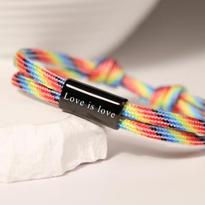 Può includere: Un braccialetto multicolore con una chiusura cilindrica nera. La chiusura presenta il testo bianco "Love is love". Il braccialetto è realizzato con un cordoncino intrecciato nei colori dell'arcobaleno, tra cui rosso, arancione, giallo, verde, blu e viola.