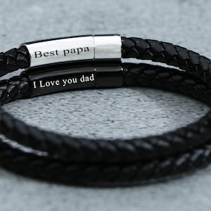 Puede incluir: Pulsera de cuero negro con dos bandas plateadas. Las bandas est&aacute;n grabadas con el texto "Best papa" y "I love you dad".