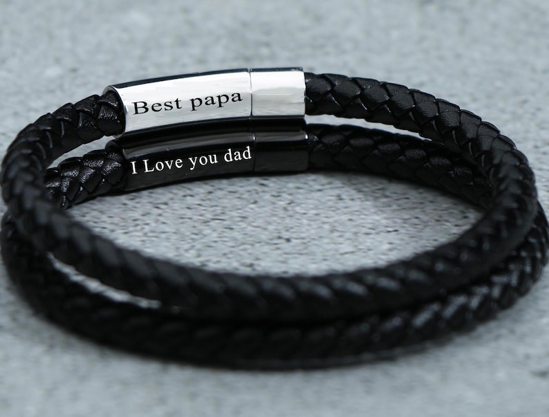 Puede incluir: Pulsera de cuero negro con dos bandas plateadas. Las bandas est&aacute;n grabadas con el texto "Best papa" y "I love you dad".