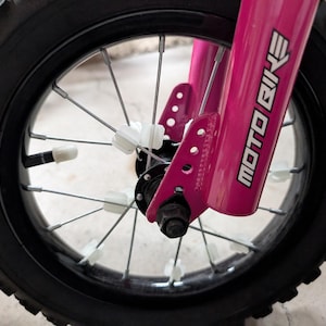 Könnte beinhalten: Nahaufnahme eines pinkfarbenen Moto Bike Vorderrads. Das Rad hat einen schwarzen Reifen mit Profil. Die Speichen sind silberfarben und die Gabel ist pinkfarben mit den Worten "MOTO BIKE" in Weiß.