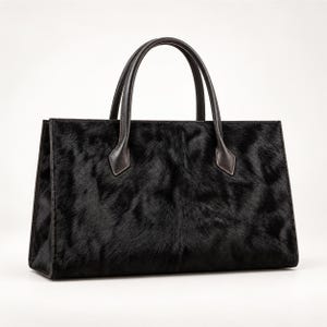 Könnte beinhalten: Eine strukturierte Handtasche mit einer schwarzen, texturierten Oberfläche aus Fell mit natürlichem Glanz. Die Tasche verfügt über zwei dunkelbraune Ledergriffe, die mit verstärkten Nähten befestigt sind, sowie einen rechteckigen Boden.