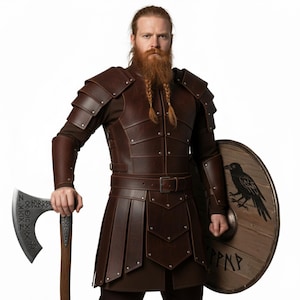 Könnte beinhalten: Braune Wikinger-Rüstung aus Leder, bestehend aus einem Panzer, Armschützern und einem plissierten Rock. Das Set ist mit Nieten und einem Gürtel versehen. Die Person hält eine Axt und einen Holzschild mit Rabenmotiv und Runen.