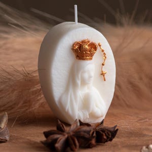Op de afbeelding: Witte ovale kaars met een gouden kroon- en rozenkransontwerp. De kaars heeft een reliëf van een religieuze figuur. Eén kaars is verpakt in doorzichtig cellofaan met een strik. Steranijs is bij de kaars geplaatst.