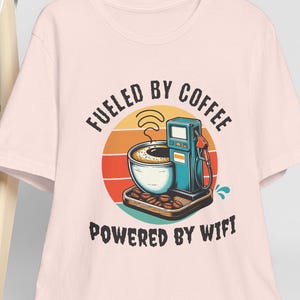 Puede incluir: Camiseta rosa claro con un diseño gráfico de taza de café y surtidor de gasolina, con el texto "Fueled by Coffee, Powered by Wifi". El diseño presenta un atardecer retro.