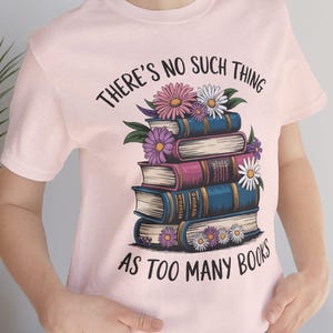 May include: A pale pink t-shirt featuring the phrase "THERE'S NO SUCH THING AS TOO MANY BOOKS" and a graphic of a stack of books decorated with flowers. The books are in shades of blue, purple, and pink.