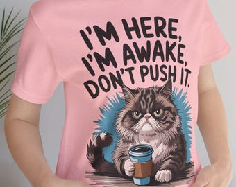 May include: Pink t-shirt with the text "I'M HERE, I'M AWAKE, DON'T PUSH IT." Features a grumpy cat holding a coffee cup. The cat is brown, gray, and white, with a blue and white background.