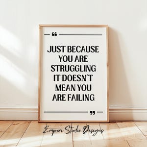 May include: Framed print with the quote "JUST BECAUSE YOU ARE STRUGGLING IT DOESN'T MEAN YOU ARE FAILING" in black text. The print is in a wooden frame and is displayed against a white wall and wooden floor.