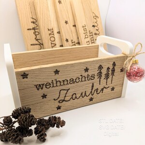 Puede incluir: Una caja de madera con temática navideña con detalles en blanco, que contiene letreros de madera con escritura. La caja está decorada con estrellas y las palabras "weihnachts Zauber". Un adorno rojo y plateado cuelga del asa.