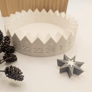 Könnte beinhalten: Ein weißer, 3D-gedruckter Adventskalenderring mit gezacktem Rand und den Zahlen 1, 22, 23 und 24. Daneben liegt ein sternförmiges, graues Ornament mit einem weißen Stern in der Mitte. Der Text "STL DATEI DIGITAL" ist sichtbar.