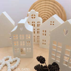 Op de afbeelding: Een verzameling witte, huisvormige kandelaars met uitgesneden ramen en deuren. Sommige hebben hartvormige uitsparingen. De huizen zijn gerangschikt op een houten oppervlak, met een houten halve cirkel op de achtergrond. Een snoer van witte kralen en dennenappels staan op de voorgrond.