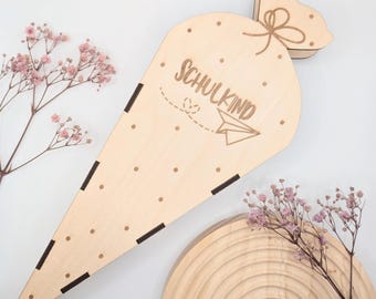 Puede incluir: Una caja de madera en forma de cono con un recorte de una bolsa en la parte superior. La caja está decorada con pequeños puntos blancos y el texto "Schulkind" con un gráfico de avión de papel. La caja está sentada sobre una superficie de madera.