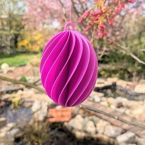 Puede incluir: Adorno esférico magenta vibrante con un diseño en espiral, suspendido de un hilo rosa. El fondo presenta una escena exterior borrosa con un estanque y árboles en flor. El texto en la parte inferior dice "SVG DATEI Digital".