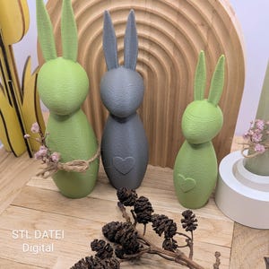Könnte beinhalten: Drei dekorative Osterhasenfiguren in Grün- und Grautönen. Die Hasen haben lange Ohren und Herzdetails. Ein Hase hat eine Schnur-Schleife. Der Hintergrund umfasst einen Holzbogen und eine Holzoberfläche.