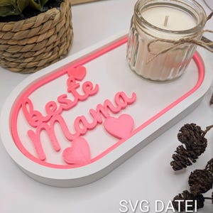 以下が含まれることがあります： ピンクの文字と2つのピンクのハートで「Beste Mama」と書かれた白とピンクの装飾トレイ。紐のリボンが付いた小さなガラス瓶のキャンドルがトレイの上にあります。小さな鉢植えの植物が背景にあります。