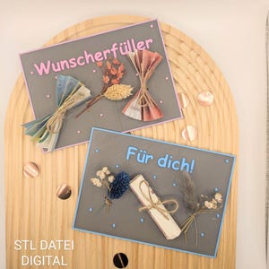 May include: Two gray rectangular cards with text and floral arrangements. One card reads "Wunscherfüller" in pink, the other reads "Für dich!" in blue. Both cards have money bundles and dried flowers. The background is a wooden surface.