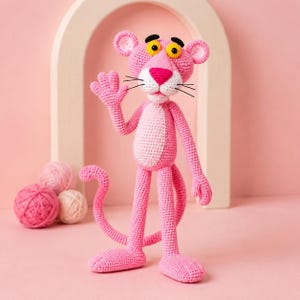 Könnte beinhalten: Eine handgefertigte Amigurumi-Häkelpuppe des Pink Panther mit einem Körper aus leuchtend rosa Garn, einem hellrosa Bauchfleck, gelben Augen mit schwarzen Pupillen und einer leuchtend rosa Nase. Die Figur hat einen langen Schwanz und eine erhobene Hand.