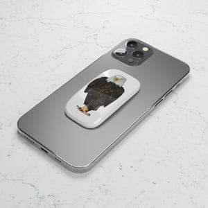 Op de afbeelding: Een witte telefoon grip met een afbeelding van een Amerikaanse zeearend. De telefoon grip is bevestigd aan een zilveren iPhone.