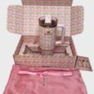 May include: A stainless steel travel mug with a handle and a clear lid, presented in a decorative box with a pink and white floral pattern. The box contains a pink satin bag and a small card. The mug has a green and white label.