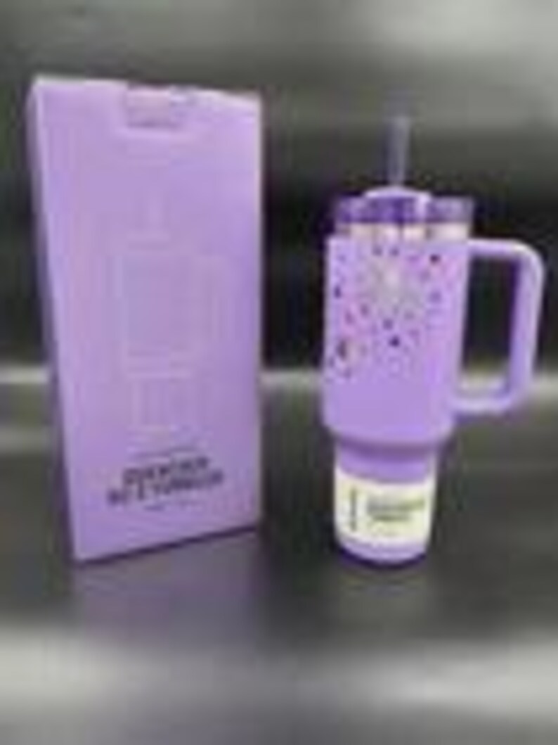 Pu&ograve; includere: Un bicchiere termico lavanda con manico e cannuccia, accanto a una scatola abbinata. Il bicchiere presenta un coperchio argentato e un design di cuori e stelle viola. La scatola ha testo e una grafica del bicchiere.