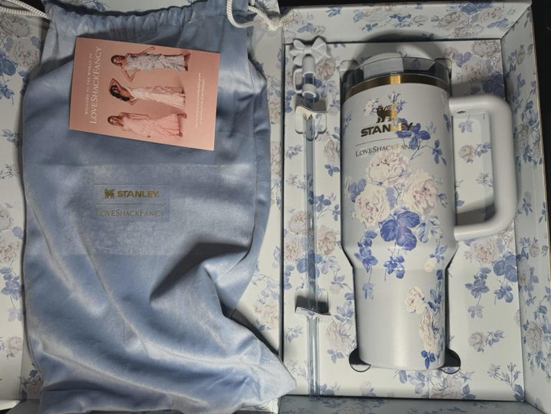 May include: A Stanley Loveshackfancy tumbler with a white handle and floral design. The tumbler is in a box with a matching floral pattern and includes a straw and a soft blue bag. The Stanley and Loveshackfancy logos are visible.