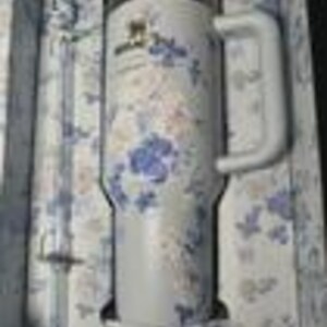 May include: A white Stanley tumbler with a handle, featuring a floral pattern in blue and gold. The tumbler is presented in a matching floral-patterned box. The tumbler has a capacity of 40 ounces.