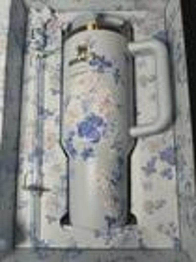 May include: A white Stanley tumbler with a handle, featuring a floral pattern in blue and gold. The tumbler is presented in a matching floral-patterned box. The tumbler has a capacity of 40 ounces.