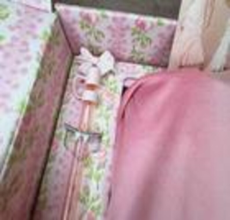 May include: A decorative perfume bottle with a white bow and a pink and gold design, presented inside a floral-patterned box. The box also contains a pink fabric. The bottle is approximately 6 inches tall.