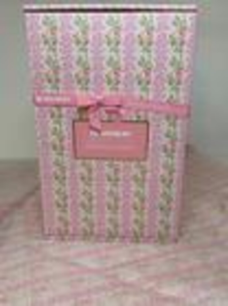May include: A rectangular gift box wrapped in pink paper with vertical stripes and a floral pattern. A pink ribbon is tied around the box, and a rectangular label is affixed to the front. The text on the label reads "The Wedding Box".