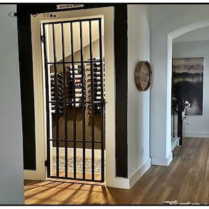 May include: A black metal gate with vertical bars, leading into a wine cellar. The cellar is filled with wine bottles in racks. The word "THE GRAPE" is above the gate. The hallway has hardwood floors and white walls.