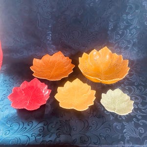 May include: A set of five ceramic bowls shaped like autumn leaves in varying sizes and colors. The collection includes one large orange bowl, one medium orange bowl, one medium red bowl, one medium yellow bowl, and one small light green bowl.