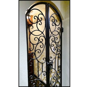 May include: Black wrought iron gate with an arched top. The gate features intricate scrollwork and a latch. The gate is set in a doorway with a light-colored wall on either side. The gate is a decorative element.