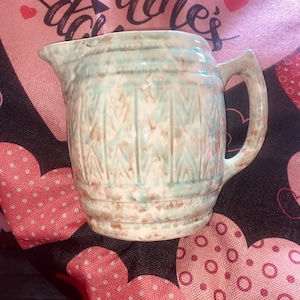 May include: A small, vintage ceramic pitcher with a handle and spout. The pitcher features a light blue and brown color scheme with a textured, patterned design. The pitcher is likely used for serving liquids.