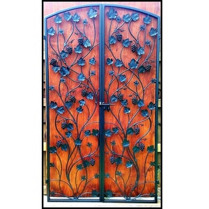 May include: A pair of wooden doors with a wrought iron design. The ironwork features grape vines with leaves and clusters of grapes. The doors are a warm brown color and have a black metal frame and hardware.