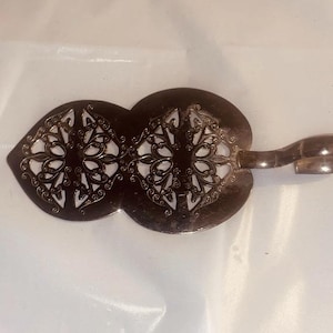 May include: A silver-toned, ornate cheese knife with a double-lobed blade featuring intricate openwork designs. The handle curves into a loop. The knife is likely used for serving cheese or other soft foods.