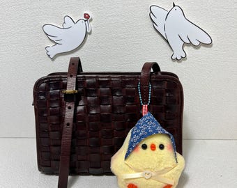 Könnte beinhalten: Eine braune, gewebte Ledertasche mit einem gelben Küken-Schlüsselanhänger. Das Küken trägt ein blaues und weiß gemustertes Bandana. Die Handtasche hat einen langen Riemen und einen oberen Griff.