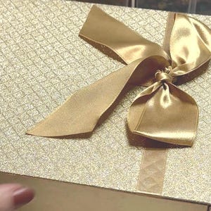 May include: A gold gift box with a large gold satin bow. The box has a glittery, textured surface.