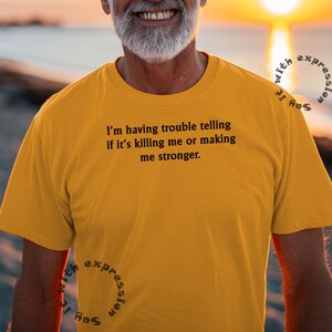 Puede incluir: Una camiseta amarilla con el texto "Estoy teniendo problemas para decir si me está matando o haciéndome más fuerte." impreso en la parte delantera.