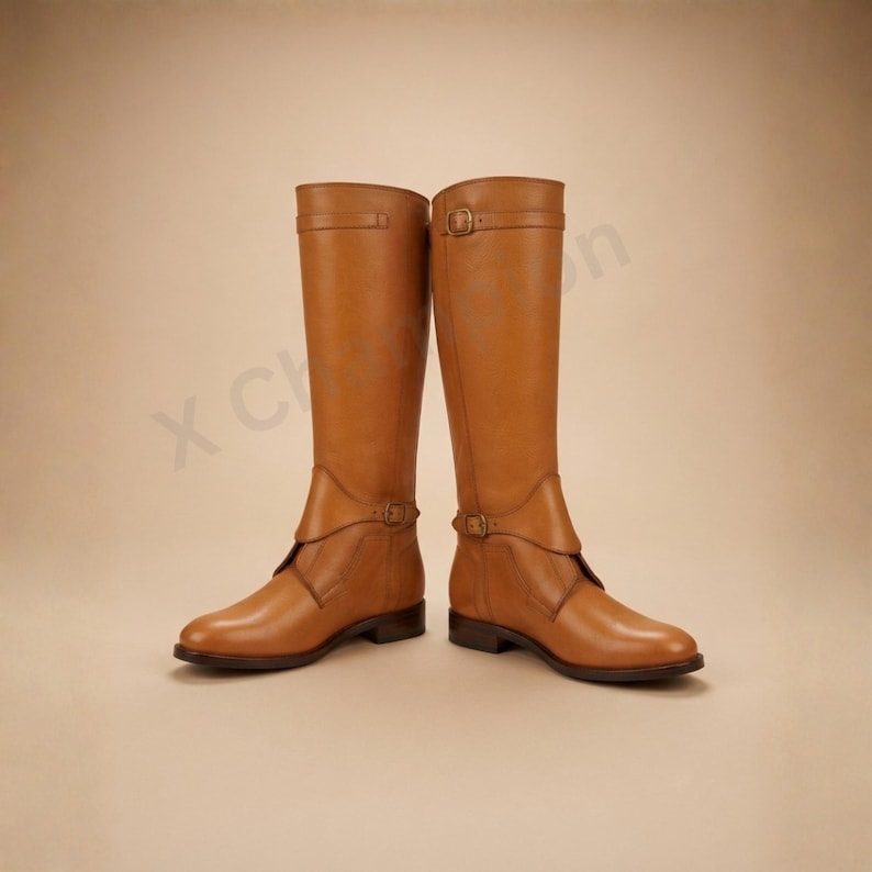 K&ouml;nnte beinhalten: Ein Paar kniehohe Stiefel aus hellbraunem Leder mit glatter Oberfl&auml;che, abgerundeter Zehenpartie und niedrigem Blockabsatz. Jeder Stiefel verf&uuml;gt &uuml;ber einen Zierriemen am Kn&ouml;chel mit Metallschnalle sowie ein passendes Riemendetail am Schaft.